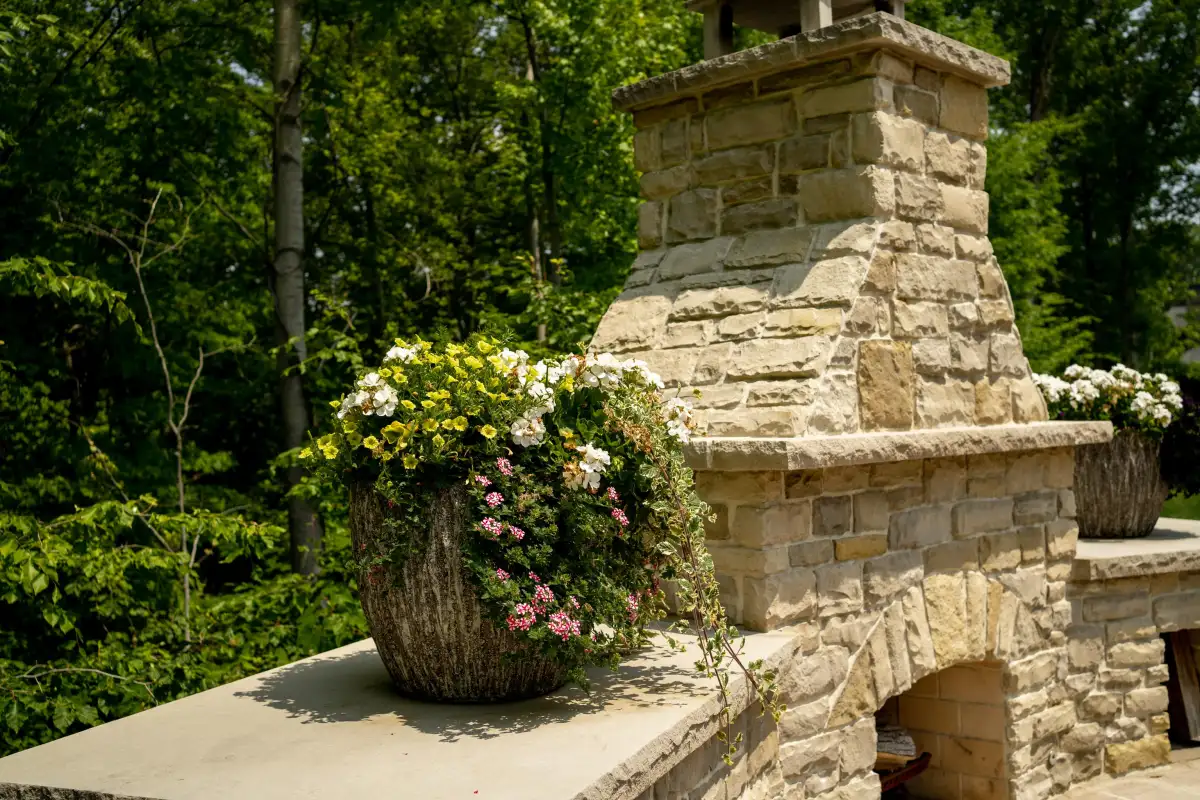Stone outdoor fireplace with overflowing flower planter on mantel in wooded backyard