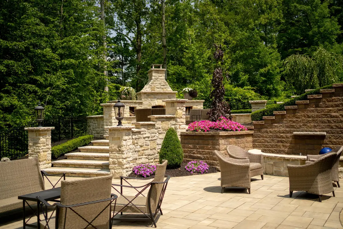 Multi-level stone patio with outdoor fireplace, wicker furniture, retaining walls, and vibrant flower beds